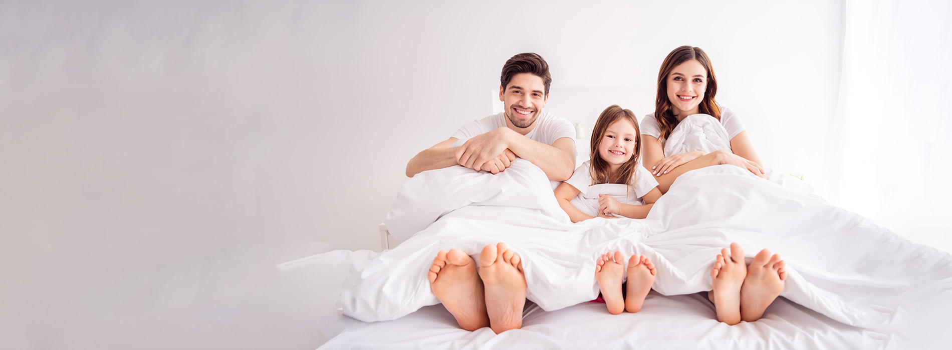 A family of four, including a man, woman, and two children, lying on a bed with a white comforter.
