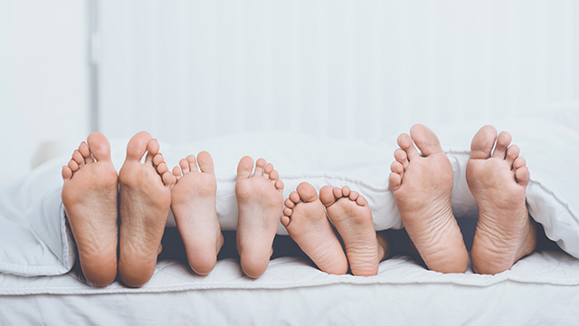 The image shows a close-up of four bare feet with toes pointing upwards, placed on a bed with a white sheet and a person lying down.