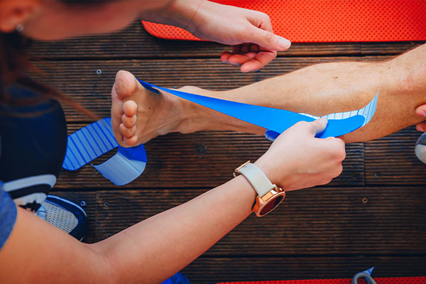 A person is taping a foot with blue tape, assisted by another individual who is holding the tape roll.